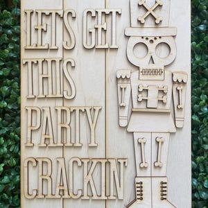 May include: Wooden sign with the phrase "LET'S GET THIS PARTY CRACKIN" and a skeleton nutcracker design. The sign is made of light-colored wood and has two holes at the top for hanging. The background is a green hedge.