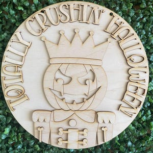 May include: A round wooden sign with the words "Totally Crushin' Halloween Queen" in a circular pattern. The sign features a carved pumpkin wearing a crown and a decorative outfit, perfect for Halloween decor.