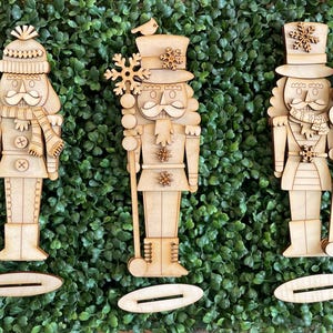 May include: Three wooden nutcracker figurines with intricate details, including hats, scarves, and snowflake accents. Each figure stands on a base, and they are set against a backdrop of green foliage. The nutcrackers are crafted from light-colored wood.