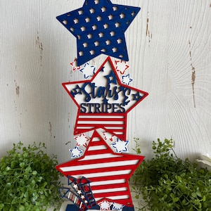 May include: A wooden patriotic star-shaped decoration with red, white, and blue stripes and stars. The decoration has the words "Stars & Stripes" written on it.