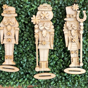 May include: Three wooden nutcracker figurines with St. Patrick's Day themes. Each figure is light brown and stands on a small base. One holds a horseshoe, another a shamrock, and all have decorative hats and details.