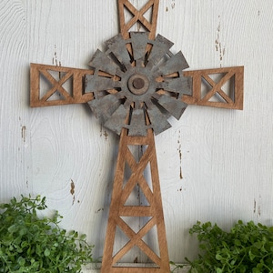 May include: A wooden cross with a metal windmill design in the center. The cross is brown and the windmill is silver with a rusty patina. The cross is on a white wooden background.