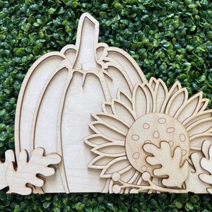 May include: Wooden cutout of a pumpkin with a sunflower and leaves. The pumpkin is outlined and has a stem. The sunflower has a center with a circle pattern. The leaves are simple and rounded.