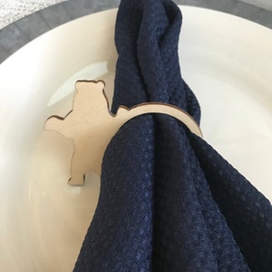 May include: A wooden bear-shaped napkin ring encircles a navy blue napkin on a white plate. The napkin ring is made of light-colored wood and features a cutout bear design. The napkin has a textured pattern.