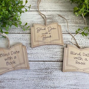 May include: Three wooden book-shaped ornaments with the text "Blind Date with a Book" on each. The ornaments are hanging from twine.