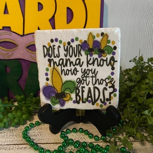 May include: White ceramic tile with a purple, green, and gold Mardi Gras design. The tile has the text "Does your mama know how you got those beads?"