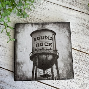 May include: Black and white photograph of a water tower with the words "Round Rock" printed on the side. The water tower is set against a cloudy sky.