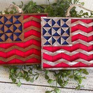 May include: Two wooden American flag ornaments with a distressed white paint finish. The flags are decorated with a geometric star pattern and a chevron pattern.