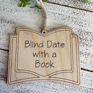 May include: Wooden ornament shaped like an open book with the text "Blind Date with a Book" engraved on it.