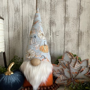 May include: A fall-themed gnome with a blue and white pumpkin patterned hat, a white beard, and an orange felt body. The gnome is sitting on a wooden surface with a blue velvet pumpkin, brown wooden beads, and a wooden leaf cutout with a similar pumpkin pattern.