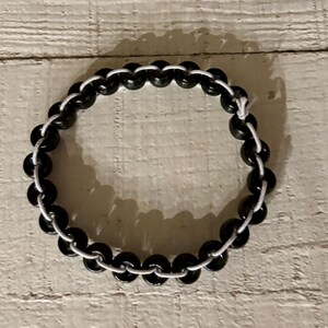 May include: A black beaded bracelet with a white cord. The bracelet is circular and made of small, round, black beads strung together with a white cord. The beads are evenly spaced, creating a simple, elegant design.