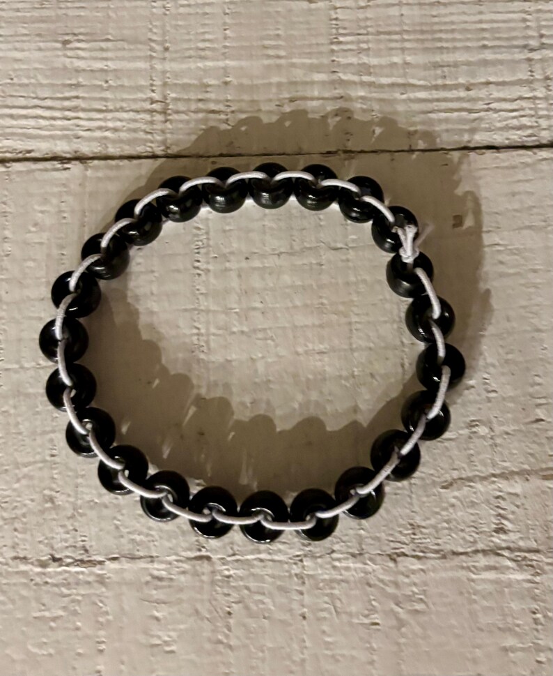 May include: A black beaded bracelet with a white cord. The bracelet is circular and made of small, round, black beads strung together with a white cord. The beads are evenly spaced, creating a simple, elegant design.
