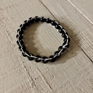 May include: A black beaded bracelet with white string. The bracelet is circular and sits on a white wooden surface. The beads are glossy and evenly spaced, creating a simple yet elegant design.