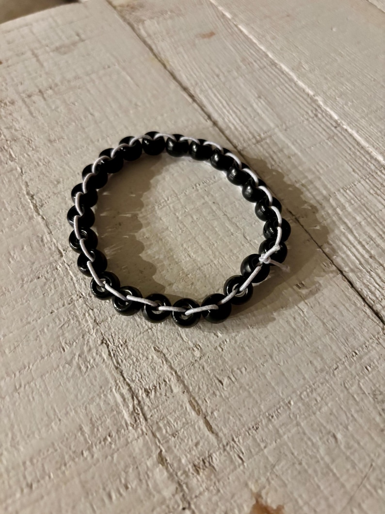 May include: A black beaded bracelet with white string. The bracelet is circular and sits on a white wooden surface. The beads are glossy and evenly spaced, creating a simple yet elegant design.