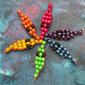 Könnte beinhalten: Ein farbenfrohes Perlenblumendesign mit sechs Blütenblättern. Jedes Blütenblatt hat eine andere Farbe, darunter Orange, Pink, Gelb, Grün, Blau und Lila. Die Perlen sind mit rosa Faden aufgefädelt, wodurch ein einzigartiges und lebendiges Stück entsteht.