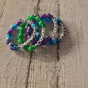 May include: Four beaded bracelets in shades of purple, green, blue, and white. Each bracelet is made of small, round beads strung together. Some bracelets have white letter beads spelling out words like "GIRL", "INTERNET", "GNARLY", and "KATSEY GAMEBOY".