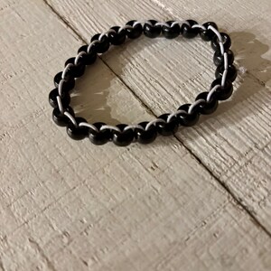 May include: A black beaded bracelet with white string woven between the beads. The bracelet is circular and sits on a light-colored, textured wooden surface. The beads are glossy and evenly spaced.