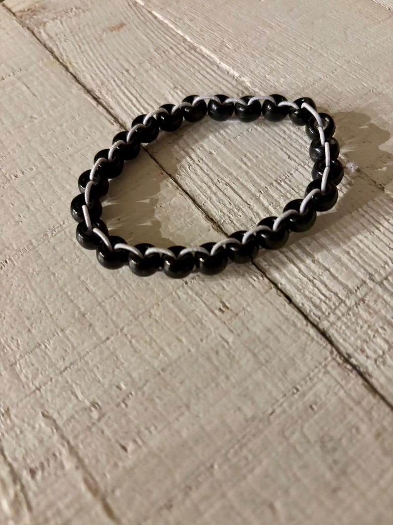 May include: A black beaded bracelet with white string woven between the beads. The bracelet is circular and sits on a light-colored, textured wooden surface. The beads are glossy and evenly spaced.