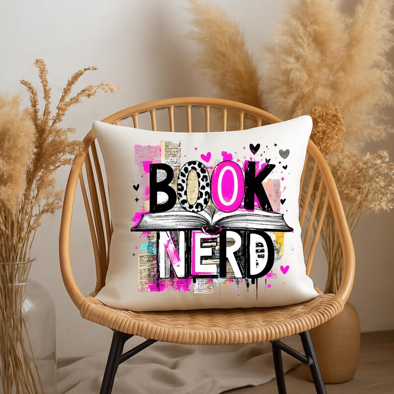 May include: A white decorative pillow with the words "BOOK NERD" in black, pink, and white. The word "BOOK" is over an open book illustration. The pillow is on a wicker chair with a neutral background.