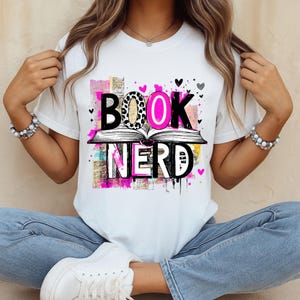 May include: White t-shirt with the words "BOOK NERD" in a colorful design. The word "BOOK" has a leopard print pattern for the letter "O". The word "NERD" is below an open book, with pink hearts scattered around the design.
