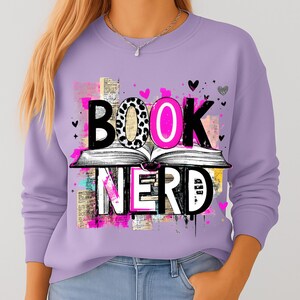 May include: A lilac-colored sweatshirt with the words "BOOK NERD" in a graphic design. The word "BOOK" is above an open book, with the letter "O" in pink and leopard print. Small hearts and abstract shapes are scattered around the design.