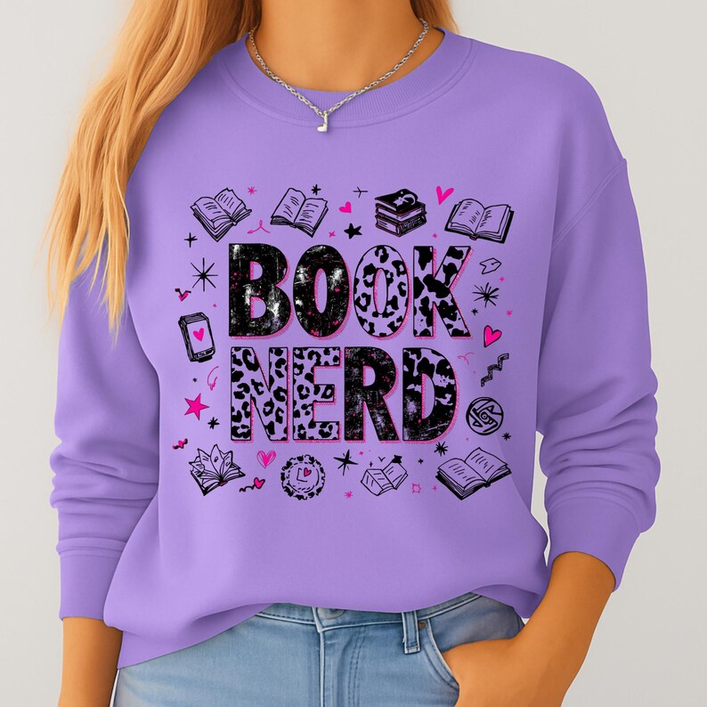 May include: A lilac-colored sweatshirt with the words "BOOK NERD" in a black and leopard print design. The sweatshirt features illustrations of books, hearts, and stars. A silver necklace is visible.