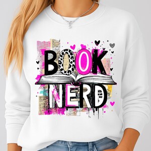 May include: White sweatshirt with a graphic that says "BOOK NERD." The word "BOOK" is above an open book, with the "O" in pink. The word "NERD" is below the book, with pink accents and black hearts.