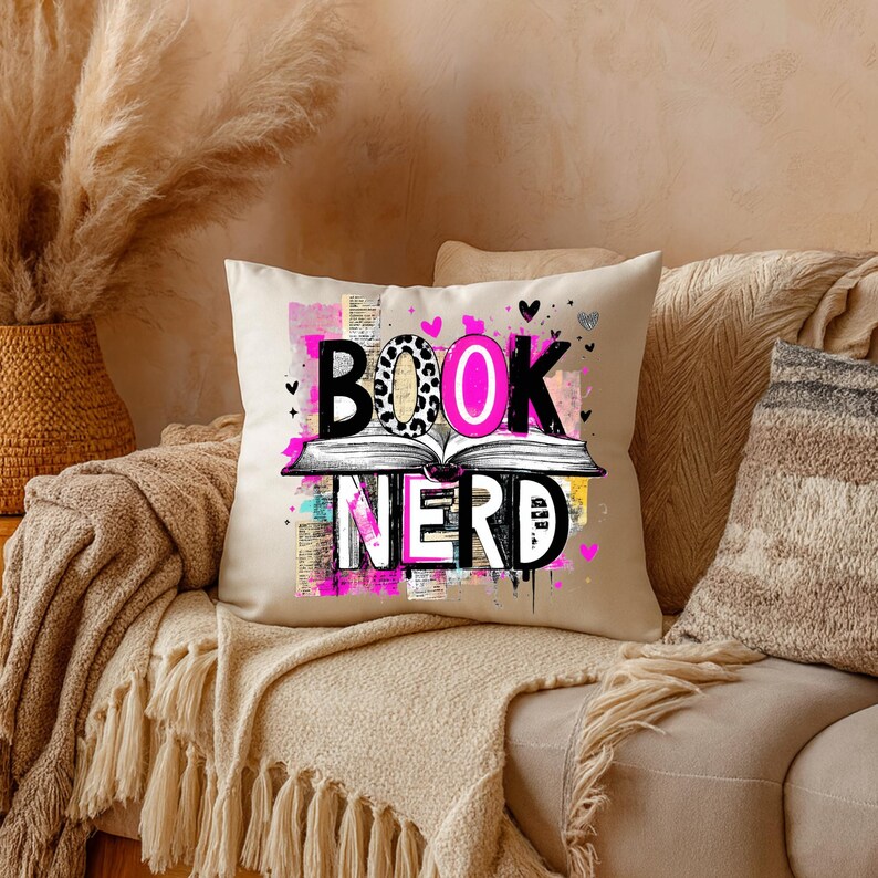 May include: A beige throw pillow with the words "BOOK NERD" in black, pink, and leopard print. The pillow features an open book graphic and small heart accents. The pillow is on a beige couch with other pillows and a fringed throw blanket.