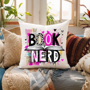 May include: A white square throw pillow with the words "BOOK NERD" in a colorful design. The word "BOOK" is above an open book illustration, with the letter "O" in a leopard print pattern. Pink hearts and accents complete the design.