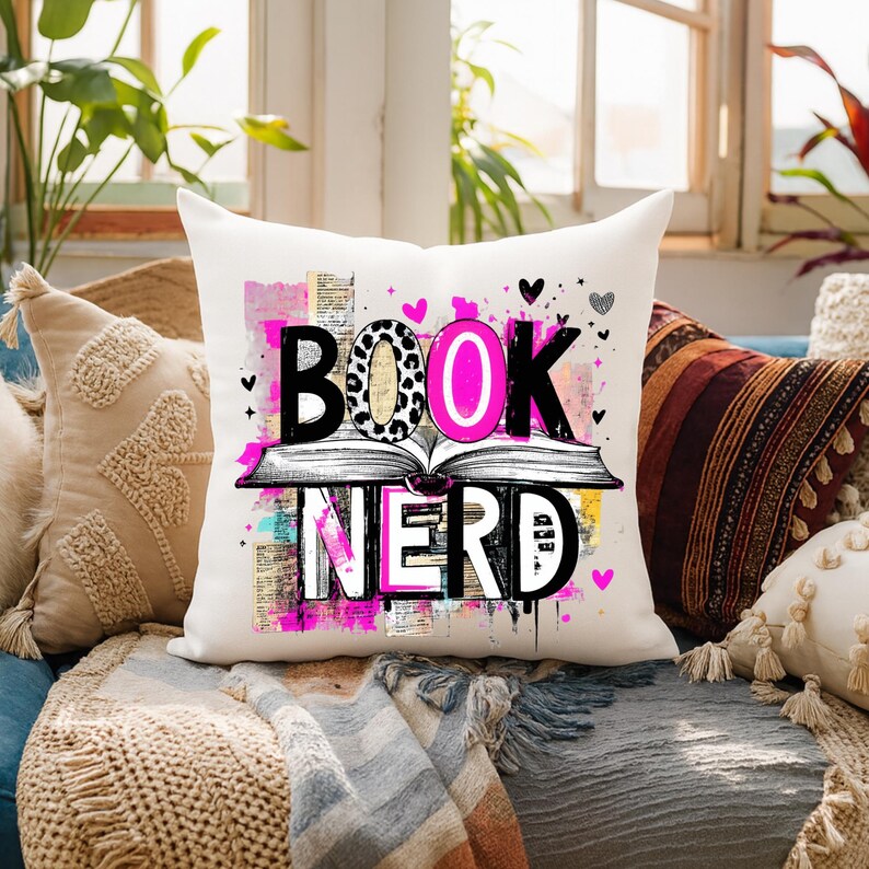 May include: A white square throw pillow with the words "BOOK NERD" in a colorful design. The word "BOOK" is above an open book illustration, with the letter "O" in a leopard print pattern. Pink hearts and accents complete the design.