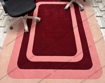 Op de afbeelding: Een rechthoekig tapijt met een gelaagd ontwerp in tinten rood en roze. Het tapijt heeft een dieprood midden, omgeven door een lichtere rode rand en een lichtroze buitenrand. Twee zwarte bureaustoelen zijn boven het tapijt geplaatst.