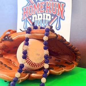 May include: A baseball-themed image showing a necklace with alternating blue and crystal-like beads draped over a baseball and mitt. A sign in the background reads "HOME RUN DRIP" with baseball bats and a baseball graphic.