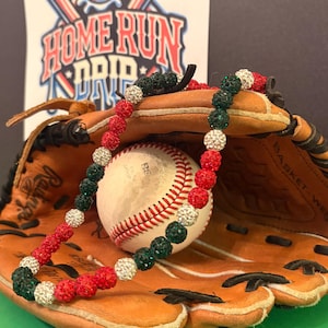 May include: A baseball necklace with red, white, and green beads rests on a baseball glove, with a baseball in the center. The background features a baseball-themed graphic with the words "HOME RUN DRIP".