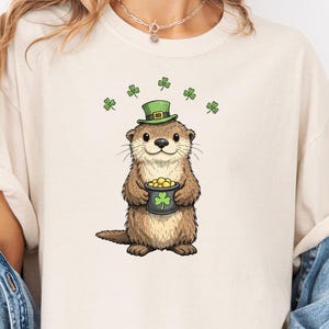 May include: Ivory-colored t-shirt featuring a cartoon otter wearing a green hat and holding a pot of gold. The otter is surrounded by green shamrocks. The shirt is a casual style.