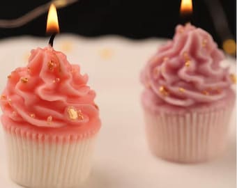 May include: Two cupcake-shaped candles with lit wicks. One candle is light pink with a darker pink frosting and gold flecks. The other candle is a darker pink with a lighter pink frosting and gold flecks. The candles sit on a white surface.