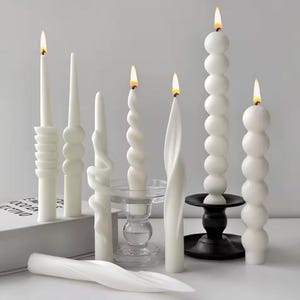 Peut inclure: Diverses bougies blanches aux formes variées, dont des spirales, des bulles et des torsades. Les bougies sont allumées, avec des flammes visibles. Certaines sont sur des supports en verre transparent et un support en métal noir.
