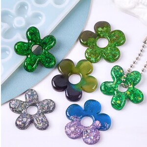 May include: A collection of flower-shaped resin pendants in various colors, including green, blue, and purple, with glitter accents. Some pendants have a hole in the center. One pendant is attached to a silver ball chain.
