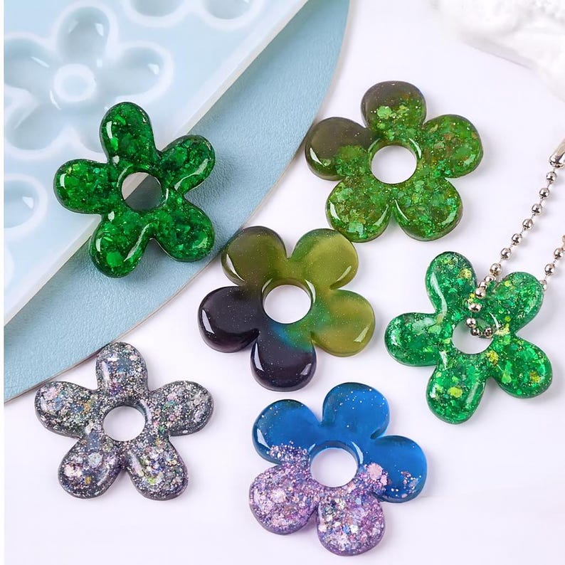 May include: A collection of flower-shaped resin pendants in various colors, including green, blue, and purple, with glitter accents. Some pendants have a hole in the center. One pendant is attached to a silver ball chain.