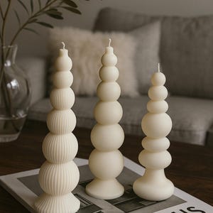May include: Three ivory-colored pillar candles with unique, stacked designs. One candle features ribbed detailing, while the others have smooth, rounded shapes. The candles are displayed on a book with the word "BAUHAUS" visible.