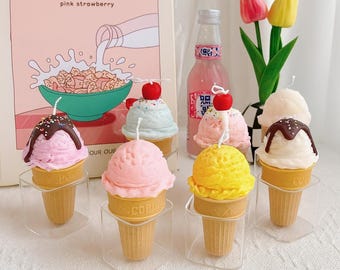 May include: A collection of ice cream cone-shaped candles in various colors, including pink, yellow, and blue, topped with a cherry. The candles sit on clear stands, with a pink strawberry-themed illustration in the background.