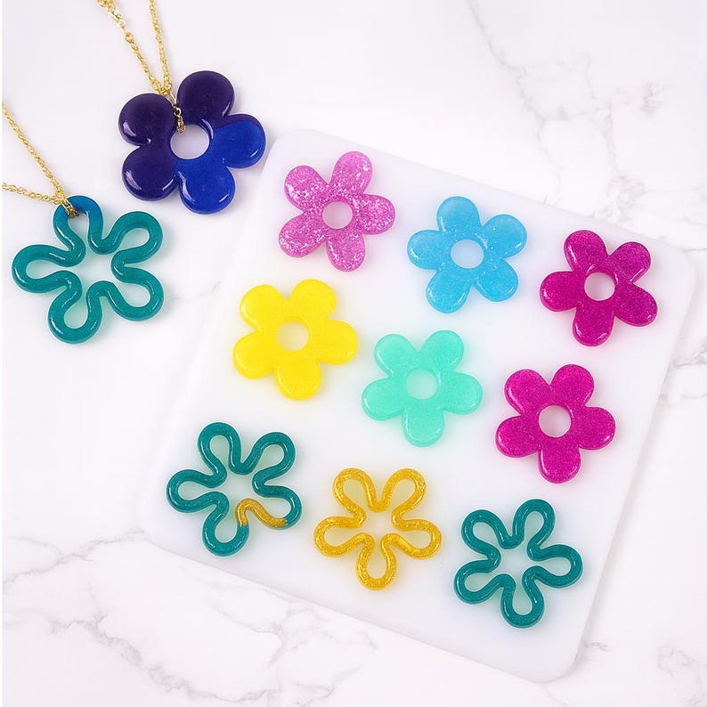 May include: A collection of colorful flower-shaped resin pendants and charms. The pendants are in shades of blue, green, yellow, pink, and gold. Two flower pendants are attached to gold necklaces. The charms are on a white surface.