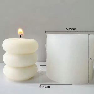 May include: A lit, ivory-colored candle with a stacked, rounded design. The candle is next to a white, cylindrical mold, 6.2 cm wide, 5.7 cm tall, and 6.4 cm deep. The candle's flame is yellow and orange.