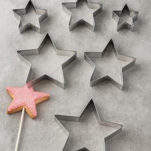 May include: A collection of star-shaped cookie cutters in various sizes, alongside a pink star-shaped cookie on a stick. The cutters are made of silver metal and are arranged on a light gray surface, suggesting a baking theme.
