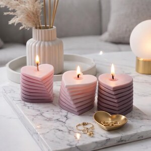 May include: Three heart-shaped candles in shades of pink and purple, with a layered design. The candles are lit, and sit on a marble tray with a gold heart-shaped dish holding jewelry. A white vase with dried flowers is in the background.