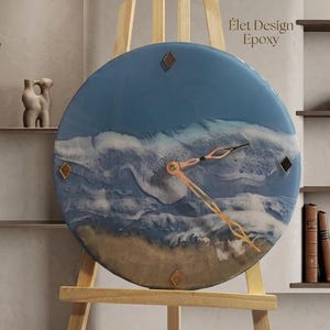 Peut inclure: Une horloge murale circulaire présentant un design en résine représentant des vagues océaniques s'écrasant sur une plage de sable. Le cadran affiche un motif de vagues bleu et blanc, une base beige, quatre marqueurs en losange, des aiguilles en or rose et le texte Élet Design Epoxy.