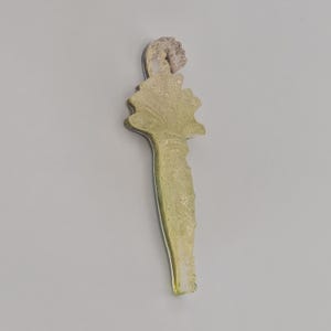 Puede incluir: Un tap&oacute;n de botella decorativo y alargado con un dise&ntilde;o texturizado verde claro. El tap&oacute;n presenta una figura marr&oacute;n claro detallada en la parte superior y una forma de hoja debajo. El tap&oacute;n est&aacute; hecho de vidrio o resina.