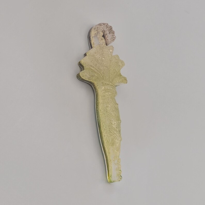 Puede incluir: Un tap&oacute;n de botella decorativo y alargado con un dise&ntilde;o texturizado verde claro. El tap&oacute;n presenta una figura marr&oacute;n claro detallada en la parte superior y una forma de hoja debajo. El tap&oacute;n est&aacute; hecho de vidrio o resina.
