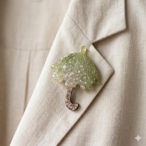 Op de afbeelding: Een groene en transparante parapluvormige broche met een gestructureerd ontwerp, bevestigd aan een beige blazer. De broche heeft een gebogen handvat en een bubbelachtige, doorschijnende uitstraling.