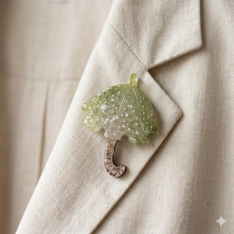 Op de afbeelding: Een groene en transparante parapluvormige broche met een gestructureerd ontwerp, bevestigd aan een beige blazer. De broche heeft een gebogen handvat en een bubbelachtige, doorschijnende uitstraling.