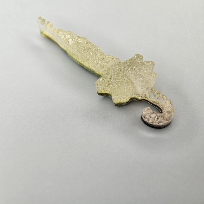 Puede incluir: Un objeto decorativo transl&uacute;cido en forma de paraguas con una superficie texturizada. El paraguas presenta un mango curvo y un dosel detallado, con una combinaci&oacute;n de colores amarillo claro y gris. El objeto es probablemente un art&iacute;culo decorativo o un amuleto.